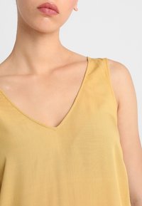 Donna che indossa un top giallo senza maniche a V, mostrando le spalle, le clavicole e parte delle labbra su uno sfondo bianco.