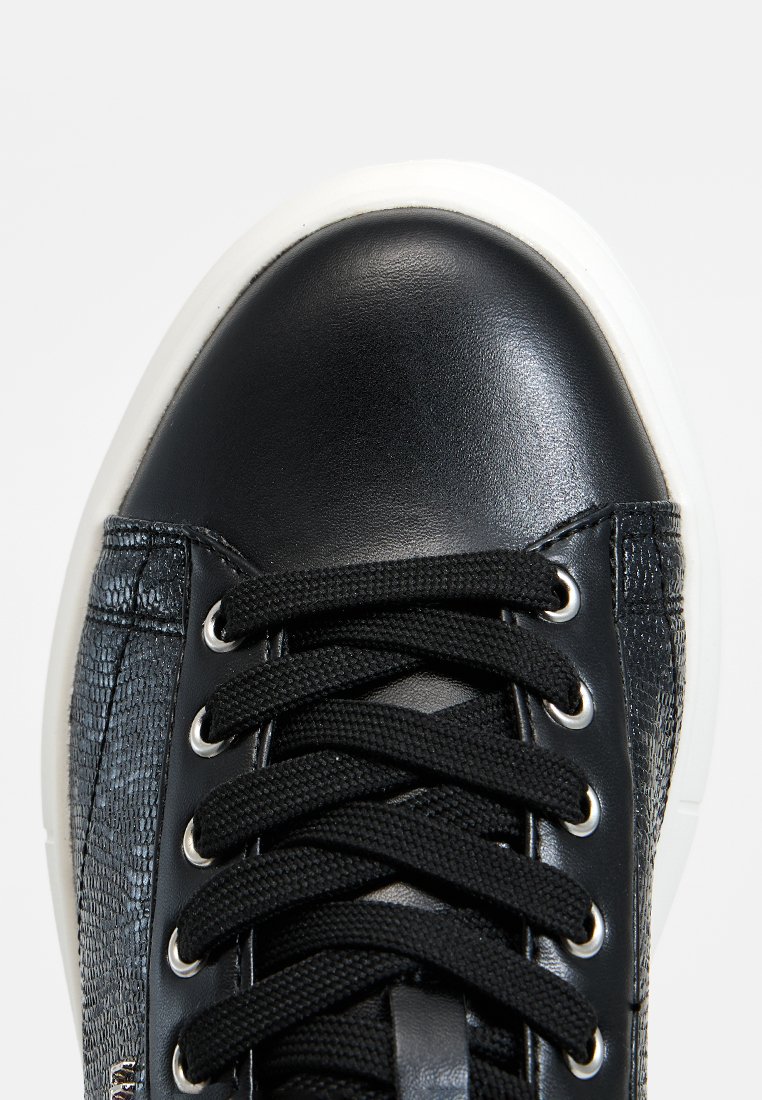 Zapatilla negra con un upper de tela negra texturizada, puntera de cuero brillante, cordones negros y suela de goma blanca. Presenta ojales plateados.