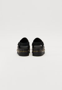 Zwarte sandalen met een dikke zool, voorzien van een klittenbandsluiting en gele stiksels langs de rand. Het materiaal lijkt duurzaam en gestructureerd.