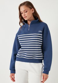 Felpa blu navy con colletto a mezzo zip, caratterizzata da strisce bianche orizzontali sul petto e polsini a costine. Tessuto morbido, vestibilità casual.
