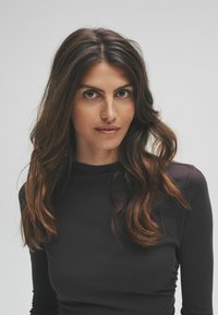 Detailný záber ženy s dlhými, vlnitými hnedými vlasmi, ktorá má na sebe priliehavý čierny rolák. Má jasnú pleť a priamy pohľad na neutrálne pozadí.