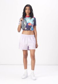 Lyhythihainen crop-top, jossa on värikäs graafinen kuvio. Laventelinväriset shortsit, joissa on kohokuvioitu kuvio. Mallilla on harmaa logo-kassi käsissään. Valkoiset lenkkarit.