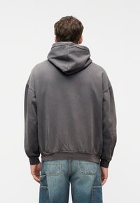 Άνδρας που φοράει ξεθωριασμένο μαύρο φούτερ με κουκούλα και ξεθωριασμένο μπλε τζιν, στέκεται με την πλάτη προς τα εμπρός σε λευκό φόντο.