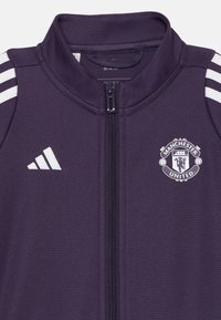 Lila zip-up jacka i texturerad tyg med vita Adidas-ränder och Manchester United-logotyp på bröstet.