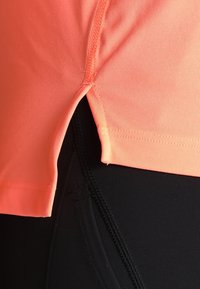 Leuchtend neonoranges Sportshirt mit seitlichem Schlitz und abgestepptem Saum, kombiniert mit schwarzen, eng anliegenden Leggings mit strukturierten Nähten.
