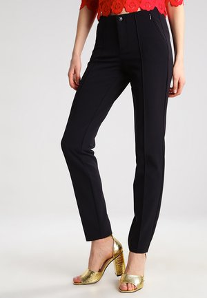 Schwarze maßgeschneiderte Hose mit glatter Textur, ausgestattet mit frontalen Reißverschlusstaschen und einem schmalen Schnitt. Kombiniert mit goldenen Sandalen mit offenen Zehen.