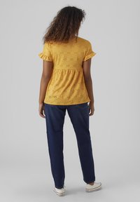 Gul bluse med broderte hull og volanger på ermene, kombinert med marineblå denimbukser. Modellen vender bort, noe som avslører detaljene i mønsteret og passformen til toppen.