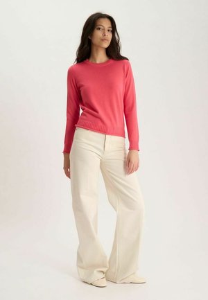 Femme debout portant un pull corail à manches longues, un pantalon large écru et des chaussures écrues sur un fond clair uni.