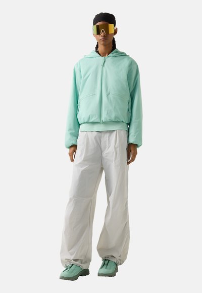 Person wearing oversized mint green hooded jacket, white wide-leg pants, mint green shoes, black headwrap, and large reflective sunglasses.