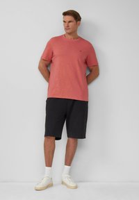 Rotes T-Shirt mit kurzen Ärmeln und Rundhalsausschnitt, kombiniert mit dunkelgrauen Shorts und weißen Sneakern. Einfaches Design, keine sichtbaren Muster oder Akzente.
