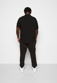 Zwarte polo met korte mouwen en een gebogen zoom, weergegeven vanaf de achterkant. Gepaard met donkere broeken en witte sneakers, met een gladde textuur.