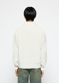 Chunky white sweater with a high collar, ribbed texture, and dropped shoulders. Worn with light blue denim, showcasing a relaxed fit.