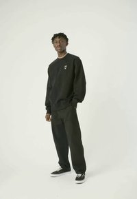 Maglione nero con logo, pantaloni neri abbinati e sneaker nere. Design semplice, vestibilità rilassata e tessuto testurizzato. In piedi su uno sfondo chiaro.