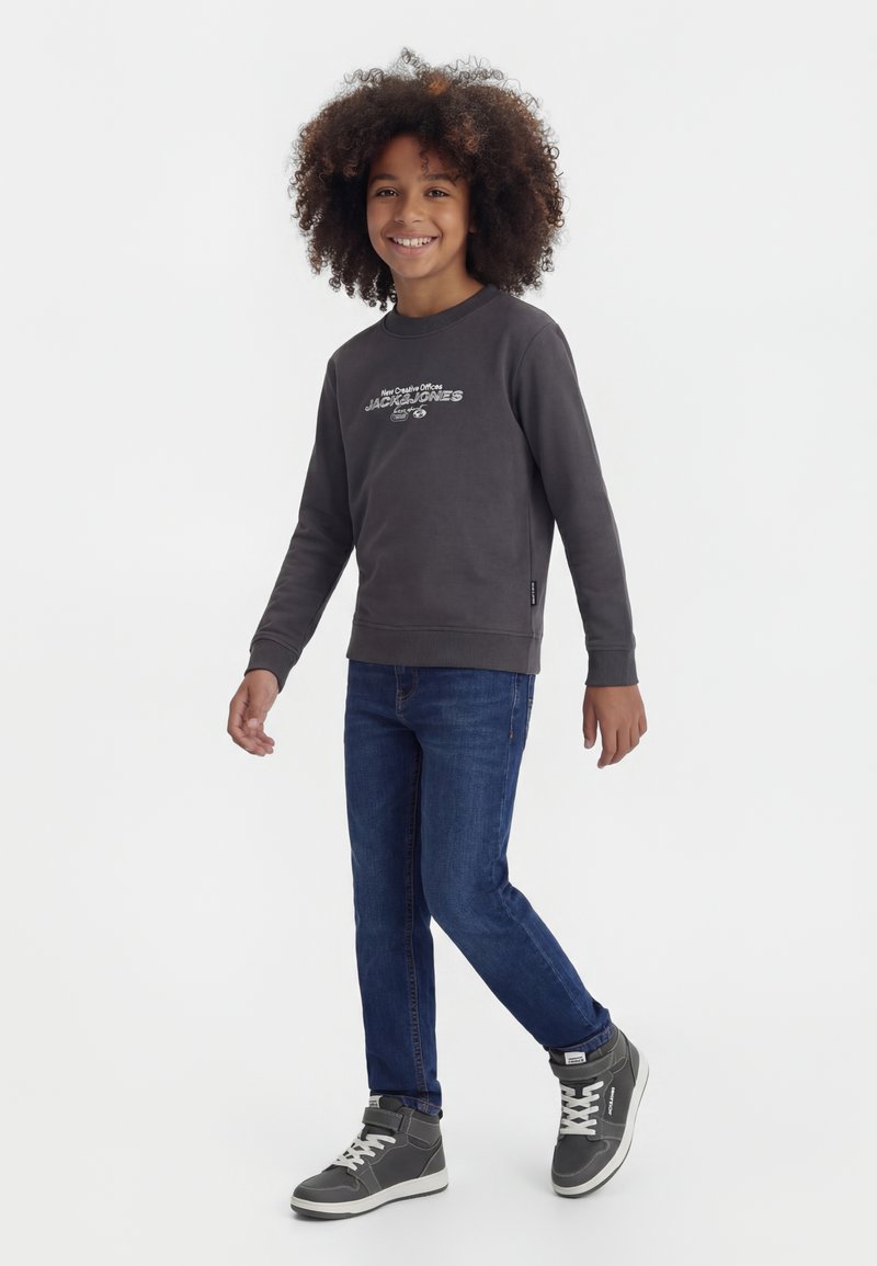 Enfant aux cheveux bouclés souriant, portant un sweat-shirt gris foncé, un jean bleu et des baskets montantes grises, marchant sur un fond blanc.