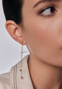 Złote kolczyki na długim wisiorku z dwoma okrągłymi koralikami zawieszonymi na cienkich łańcuszkach, przymocowane do małego wkrętu. Czysty, nowoczesny design.