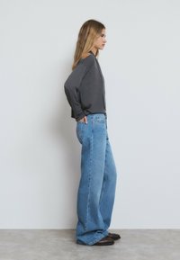 Un haut gris à manches longues et un jean large bleu clair. Tissu texturé avec une coupe décontractée. Le modèle se tient devant un fond blanc uni.