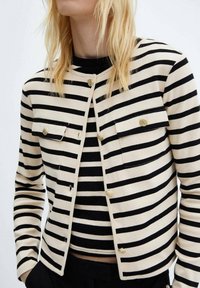 Pasiasta kurtka w kolorze kremowym i czarnym, z dwoma kieszeniami na piersi i złotymi elementami. Wykonana z miękkiego materiału, z okrągłym dekoltem i zapięciem na guziki.