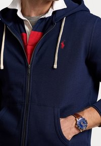 Marinblå zip-up hoodie med vita dragband och en liten röd logga. Lagrad över en röd och grå kragskjorta. Brun armbandsur på handen.