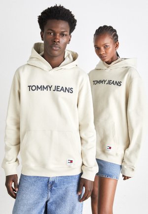 Sudaderas beige con "TOMMY JEANS" en letras oscuras, bolsillos frontales tipo canguro, puños acanalados y parche con el logo en el dobladillo; combinadas con pantalones cortos de mezclilla.