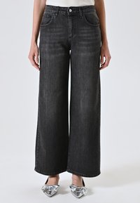 Persoon in hoog-gesneden, wijd uitlopende zwarte jeans en zilveren, puntige ballerina's met grote strikken, staand voor een effen achtergrond.