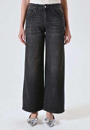 Person wearing high-waisted wide-leg black jeans and silver pointed-toe flats with large bows, standing against plain background.