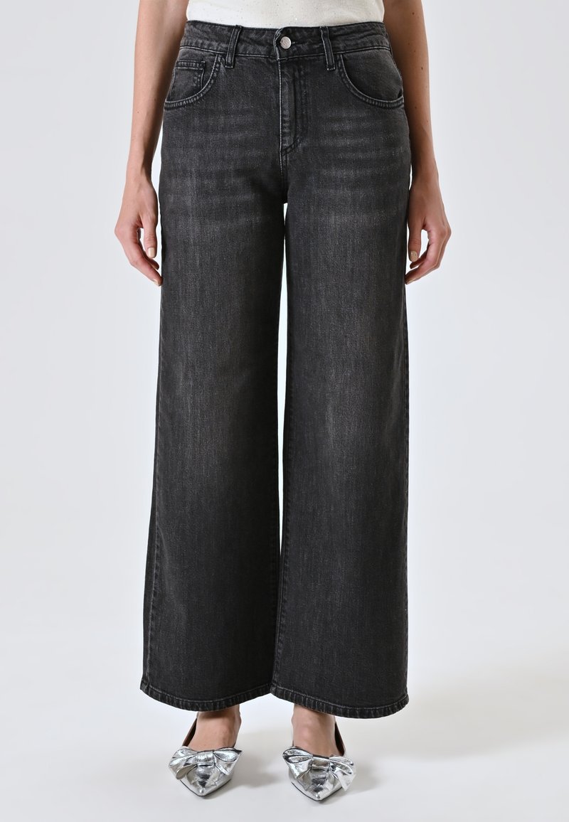 Persoon in hoog-gesneden, wijd uitlopende zwarte jeans en zilveren, puntige ballerina's met grote strikken, staand voor een effen achtergrond.