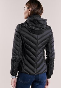Giacca a vento nera con design a motivo chevron, cappuccio removibile, chiusura con zip e polsini a coste. Indossata con jeans blu.