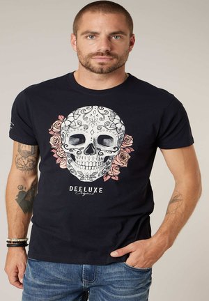 Man met een zwart T-shirt met een decoratieve witte schedel en roze rozen, een blue jeans en meerdere tatoeages op beide armen.