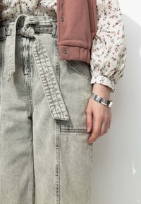 Pantalons en denim gris clair taille haute avec une ceinture nouée, d'une coupe ample et dotés de poches latérales. Portés avec un blouse à manches longues à motif floral et un bracelet en argent.