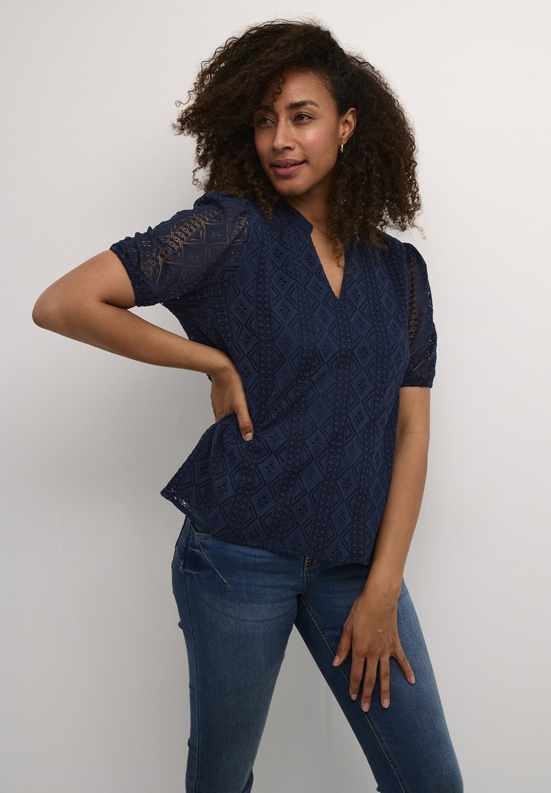Blusa in pizzo blu navy con motivi geometrici, maniche a puff corte e design a scollo a V, abbinata a jeans in denim.