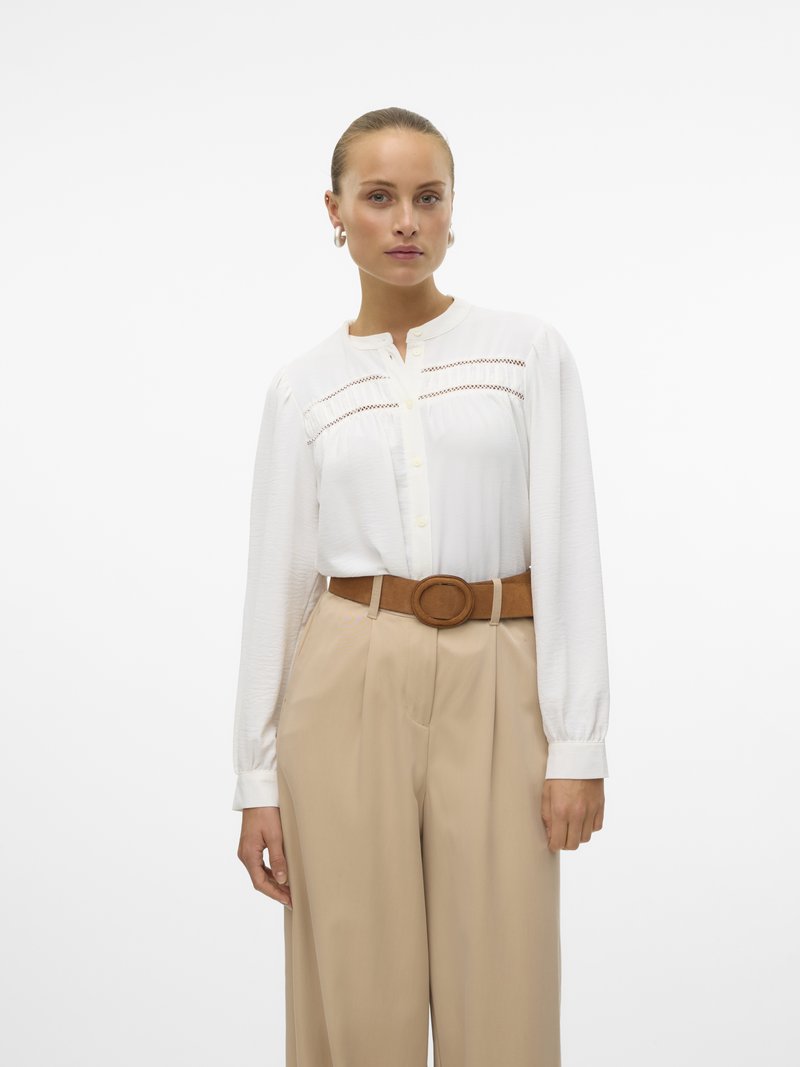 Femme aux cheveux plaqués en arrière porte un chemisier blanc à manches longues avec des détails en dentelle, un pantalon large beige taille haute et une ceinture marron à boucle ronde.