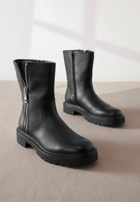 Bottines en cuir noir avec fermeture éclair sur le côté, semelle en caoutchouc épaisse et texture lisse. Présente des coutures discrètes et un design à bout rond.