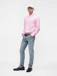 Homme portant une casquette blanche, une chemise rayée rose à boutons, un jean bleu clair et des mocassins noirs, debout les mains dans les poches, fond neutre.