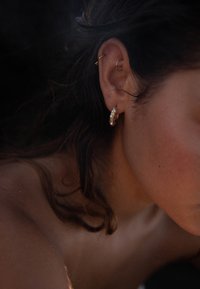 Guldörhängen med pärlaccent och en smal hoop-piercing, mot en bakgrund av löst, vågigt brunt hår och en mjuk hudtextur.