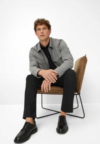 Grijze button-up jas over een zwart shirt, zwarte broek en zwarte schoenen. De stoel heeft een bruine stoffen kussen met een minimalistisch metalen frame.