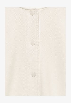 Detail bílého texturovaného látky s vertikálním řádkem kulatých patentek ve stejné barvě.