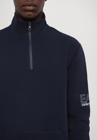 Sudadera con cremallera de color azul marino y cuello alto, con un bolsillo frontal y un logo bordado en blanco en la manga. Tela suave y texturizada.