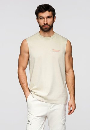 Brodaty mężczyzna o ciemnych włosach, ubrany w beżowy bezrękawnik i białe spodnie typu cargo, stojący z jedną ręką w kieszeni na jednolitym tle.