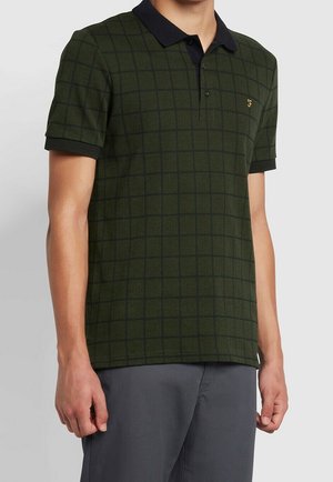 Uomo che indossa una polo a maniche corte verde scuro con colletto nero e motivo a griglia, abbinata a pantaloni grigio scuro.