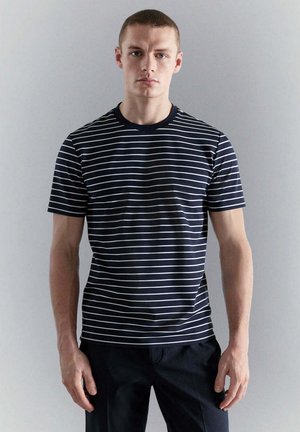 Joven con el pelo corto que lleva una camiseta de rayas horizontales azul marino y blanca y pantalones oscuros, de pie contra un fondo claro y liso.