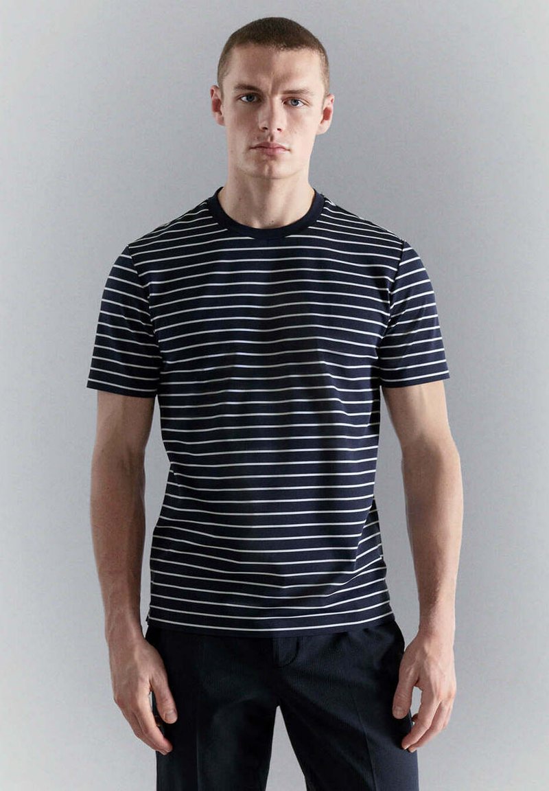 Jeune homme aux cheveux courts portant un t-shirt rayé horizontal bleu marine et blanc et un pantalon foncé, debout sur un fond clair uni.