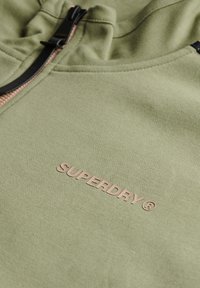 Sudadera verde hecha de un tejido suave y texturizado, con una cremallera negra y un logo en relieve de "SUPERDRY" en rosa claro.