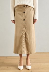 Jupe midi en coton kaki avec fermeture à boutons à l'avant et deux poches latérales. Texture lisse et coupe droite. Portée avec des talons hauts blancs.