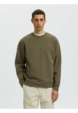 Man met kort donker haar, die een olijfgroene trui en beige broek draagt, staat voor een effen lichte achtergrond.