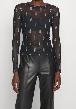 Black sheer long-sleeve top with repeated white "HUGO" logos worn over a black bandeau, paired with high-waist black leather pants.