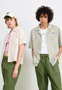 Deux personnes portent des tenues décontractées : l'une porte une chemise courte à rayures beiges et un pantalon vert ; l'autre est en chemise vert clair et pantalon assorti.