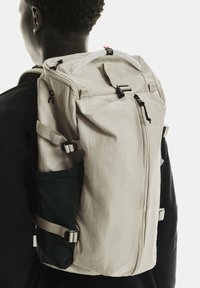 Personne portant un sac à dos beige avec une poche latérale noire et plusieurs sangles ajustables sur des vêtements noirs, montré de dos et de côté.