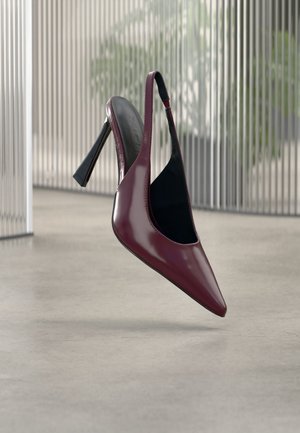 Slingback lucido di colore rosso scuro con tacco alto, punta appuntita e fodera interna nera, che fluttua sopra un pavimento in cemento liscio all'interno.