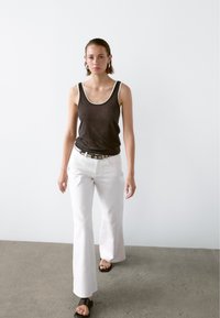 Černý tílko s šedým lemem, bílé rozšířené kalhoty a černé sandály. Modelka stojí na betonové podlaze s minimalistickým pozadím.