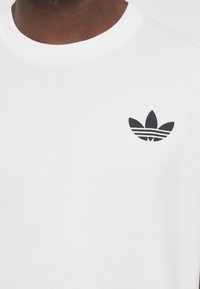 Hvid bomulds T-shirt med et sort tre-blads logo på venstre brystområde. Stoffet ser glat og let ud.